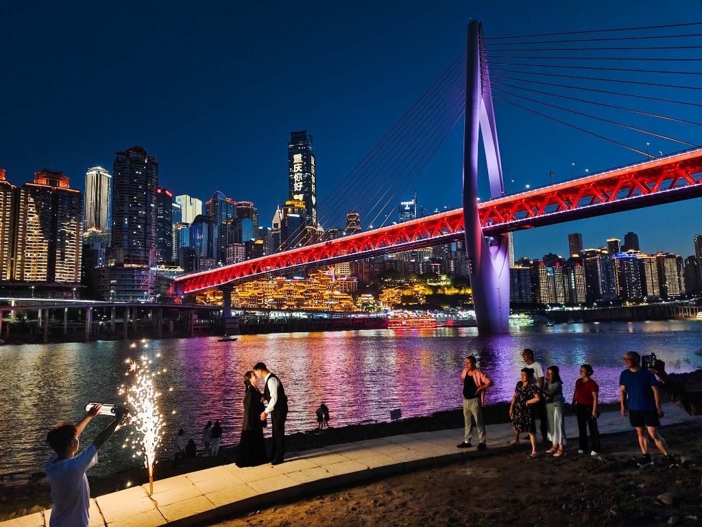 2022年4月26日，重庆市江北嘴嘉陵江边，市民以夜幕中的渝中区为背景拍摄照片，旁边一群市民用微笑围观表示祝福。